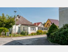 Straßenszene in einer Wohngegend mit kleinen Einfamilienhäusern, eingefriedet von niedrigen Zäunen, und Vorgärten mit Blumen und Sträuchern. Im Hintergrund sind weitere Häuser mit Satteldächern zu sehen, und ein Schild mit der Aufschrift „Uferstraße“ steht an einer Straßenlaterne.