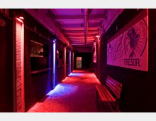 Ein schmaler Flur mit Betonboden und industriellem Design, beleuchtet von roten und blauen Neonlichtern. An den Wänden hängen Poster, auf einem steht „TRESOR.“, und rechts steht eine Holzbank.