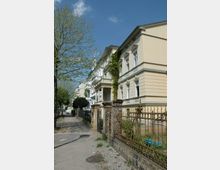 Reihe von historischen Wohnhäusern mit hellem Stuck und Verzierungen, teils bewachsen mit Efeu; im Vordergrund ein schmiedeeiserner Zaun und ein gepflasterter Gehweg mit Bäumen.