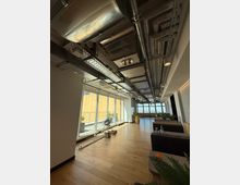 Modern office lounge corridor with light wood flooring, large floor-to-ceiling windows along one side, and an exposed industrial ceiling with visible ductwork, pipes, and cable trays. Low seating and planters line the right side, with a balcony railing and city view visible through the windows.