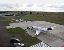 Kleiner Flughafen mit einem geparkten Privatjet auf einer asphaltierten Fläche. Im Hintergrund sind Hangars, Tankanlagen und eine weitläufige Wiesenlandschaft sichtbar.