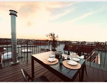 Eine Dachterrasse mit einem Esstisch und vier Stühlen, gedeckt mit Tellern, Gläsern und Flaschen, im Vordergrund. Der Boden besteht aus Holzplanken, und ein Geländer mit Metallstreben bietet Ausblick auf umliegende Dächer. Im Hintergrund ist der Himmel während des Sonnenuntergangs zu sehen.