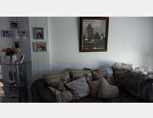 Gemütliches Wohnzimmer mit dunklem Sofa und vielen Kissen vor einer weißen Wand, darüber ein gerahmtes Landschaftsgemälde. Links steht ein gläsernes Eckregal mit Vase und Blumen vor einer vertäfelten Wand mit mehreren gerahmten Fotos.