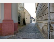 Schmale Gasse mit gepflastertem Boden, flankiert von einer rosa-gestrichenen Fassade links und einer beige-grauen, teilweise beschädigten Wand rechts. Im Hintergrund ein modernes Gebäude mit Glasfront und ein kleineres, helles Haus mit Satteldach.