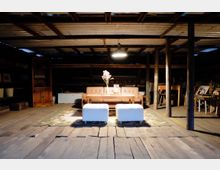 Gemütlicher Sitzbereich in einem rustikalen Dachboden mit Holzfußboden und Wellblechdecke. Zentrales Möbelstück ist ein braunes Ledersofa, flankiert von einem Holztisch und zwei weißen Hockern, auf einem Teppich platziert, umgeben von alten Möbeln und Dekorationen.