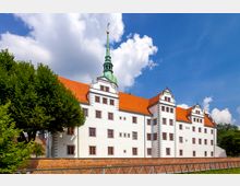 Ein historisches Schloss mit weißer Fassade, roten Dachziegeln und einem grünen Kirchturm mit vergoldeter Spitze. Vor dem Gebäude befinden sich eine Ziegelsteinmauer und Bäume, dahinter ein klarer blauer Himmel mit einigen Wolken.