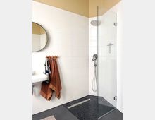 Modern bathroom with white tiled walls, a walk-in shower enclosed by a clear glass panel, and a pebble-stone shower floor with a linear drain. A wall-mounted sink, round mirror, and a towel rack with hanging towels sit beside the shower.