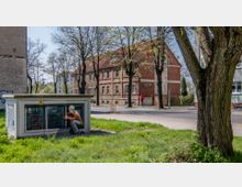Straßenszene mit einem gemauerten Gebäude mit rotem Ziegeldach im Hintergrund, auf dem "ANTIK" geschrieben steht. Im Vordergrund befindet sich eine Stromverteilerbox mit der Bemalung eines Bauarbeiters, umgeben von Grünflächen und Bäumen.