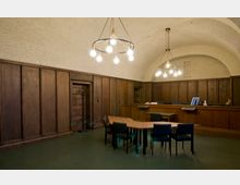 Innenraum eines klassischen Verwaltungs- oder Versammlungsraums mit Holztäfelung an den Wänden, einer Holztür und einer Empfangstheke. Der Raum hat eine gewölbte Decke, schlichte Leuchter mit Glühbirnen und einen Tisch mit mehreren Stühlen im Vordergrund.