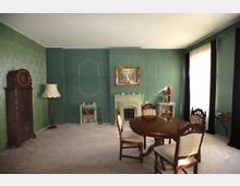 Wohnzimmer mit grün gestrichenen Wänden und klassischen Verzierungen, einem Kamin in der Mitte, einem runden Holztisch mit gepolsterten Stühlen, einer altmodischen Standuhr und einem beigefarbenen Teppichboden. Große Fenster mit dunklen Vorhängen lassen Tageslicht einfallen.