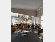 Heller Raum eines Cafés mit moderner Einrichtung, darunter schwarze Tische, Holzstühle und eine orangefarbene Couch. Die Wände sind minimalistisch dekoriert mit Bildern und hängenden Objekten, während die Nachmittagssonne durch große Fenster Schatten im Raum wirft.