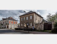 Straßenszene mit zwei zweistöckigen Gebäuden: rechts ein schlichtes Wohngebäude mit abgenutzter Fassade und angrenzendem Tor, links ein historisches Eckgebäude mit roten Klinkerelementen und dekorativen Fensterrahmen. Im Vordergrund führt ein Gehweg entlang, und ein Straßenlaternenmast steht zentral im Bild.