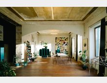 Loftartiger Raum mit Holzboden und abgenutzten Decken, dekoriert mit farbenfrohen Wandgemälden und Pflanzen. An den Wänden befinden sich Regale und Kunstwerke, Tageslicht fällt durch große Fenster mit schwarzen Vorhängen.