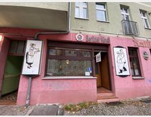 Außenansicht eines Restaurants in einem Wohngebäude mit rosa Putzfassade und grünen Wänden. Über der Tür und dem Fenster steht "Berliner Kindl", daneben befinden sich humorvolle Zeichnungen eines Kellners und einer Bedienung.