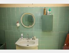 Badezimmer mit blassgrünen Wandfliesen, einem weißen Waschbecken mit Einhebelmischer, rundem Wandspiegel und einem kleinen Hängeschrank. Auf dem Waschbecken und Schrank stehen Pflegeprodukte, daneben hängt ein Zahnputzbecher mit Zahnbürsten.