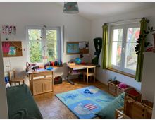 Helles Kinderzimmer mit zwei Fenstern, einem Schreibtisch und einer Sitzbank. Auf dem Boden liegt ein bunter Teppich mit Meeresmotiven, daneben befinden sich Körbe und Spielzeug. An den Wänden hängen Bilder und Karten, die Einrichtung ist überwiegend aus Holz.