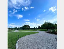 Weitläufiger Park mit geschwungenen Kieswegen, umgeben von grünen Rasenflächen, einzelnen Bäumen und Blumenbeeten; im Hintergrund ein blauer Himmel mit weißen Wolken.