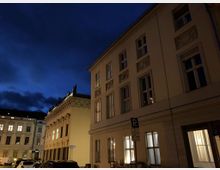 Straßenszene bei Nacht mit historischen Gebäuden, die beige Fassaden und hohe Fenster haben. Im Vordergrund sind parkende Autos und ein Parkplatzschild zu sehen, während der Himmel dunkelblau mit Wolken ist.