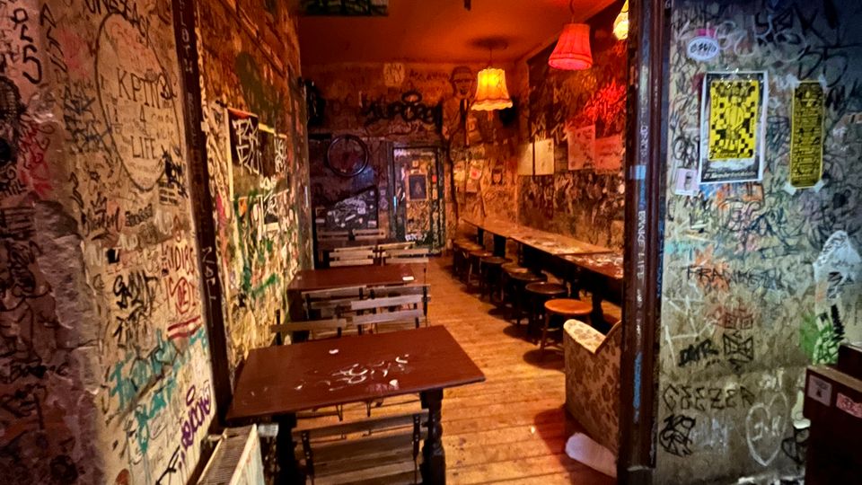 Schmaler Bar- oder Kneipeninnenraum mit Holzfußboden, kleinen Tischen und Bänken sowie einer langen Sitzbank mit Hockern entlang der rechten Wand. Wände, Decke und Türen sind dicht mit Graffiti und Aufklebern bedeckt, beleuchtet von warmen, hängenden Lampenschirmen.