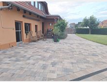 Hofbereich eines Hauses mit orangefarbener Fassade und rotem Ziegeldach, gepflastertem Boden sowie Rattanmöbeln. Im Hintergrund befinden sich ein Zaun, Büsche, ein Baum und angrenzende Gebäude.