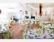 Festlich dekorierter Saal mit runden Tischen, die mit weißen Tischdecken, Geschirr und kleinen Blumengestecken geschmückt sind. Über den Holzboden sind Ballons, weiße Stoffbahnen an der Decke sowie eine zentrale Dessert- und Getränketafel erkennbar.