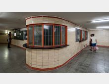 Ansicht eines U-Bahn- oder Bahnhofszugangs mit beige gefliesten Wänden, roten Fensterrahmen und einem halbrunden Schalterbereich. Personen stehen vor Wandtafeln und einer Ticketstelle, während der Boden aus grauen Fliesen besteht.