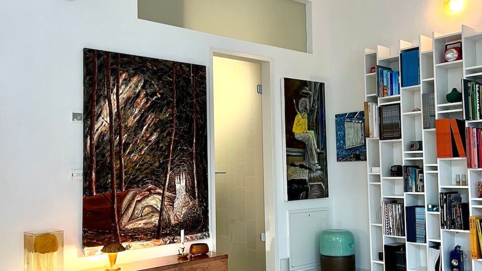 Bright, modern room with white walls and polished light floor, featuring exposed ceiling beams and warm accent lighting. Large abstract paintings hang beside a doorway, and a tall white shelving unit filled with books and objects stands along the right wall.