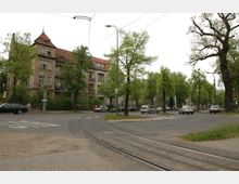 Mehrstöckiges Wohngebäude mit roten Ziegeldächern und verputzter Fassade an einer Baumallee, im Vordergrund eine Straßenkreuzung mit Straßenbahnschienen und vorbeifahrenden Autos. Der Bereich ist von großen, grünen Bäumen gesäumt.