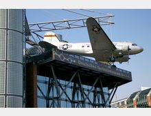 Ein silbernes Flugzeug (Douglas C-47) ist an einer erhöhten Stahlkonstruktion außerhalb des Deutschen Technikmuseums in Berlin angebracht. Im Vordergrund ist ein moderner Gebäudeteil mit Glas- und Ziegelarchitektur sowie einem Schriftzug des Museums sichtbar.