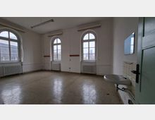 Leerer, schlicht gehaltener Innenraum mit hohen weißen Wänden, drei großen Rundbogenfenstern und Heizkörpern darunter, beleuchtet durch Deckenleuchten. Rechts sind eine Tür und ein wandmontiertes Waschbecken mit Fliesenrückwand zu sehen.