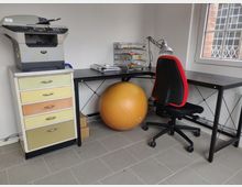 Arbeitsbereich mit einem schwarzen Schreibtisch, einem schwarzen Bürostuhl mit rotem Rand, einem gelben Gymnastikball und einem Drucker auf einer bunten Kommode. Im Hintergrund steht ein Fenster mit Gitter und Blick auf eine Ziegelwand.