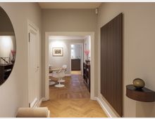 Ein eleganter Flur mit Holzparkettboden, cremefarbenen Wänden und einer modernen Einrichtung. Im Hintergrund befindet sich ein Essbereich mit weißen Stühlen, einem Holztisch und dekorativen Elementen wie einer roten Vase und einem gerahmten Kunstwerk an der Wand.