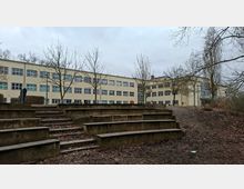 Großer, dreigeschossiger Schul- oder Verwaltungsbau mit heller Fassade und vielen rechteckigen Fenstern, im Hintergrund eines Innenhofs. Im Vordergrund stehen terrassenförmige Betonstufen wie eine kleine Freilufttribüne, umgeben von kahlen Bäumen und unbefestigtem Boden.