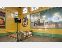 Gelb gefliester U-Bahnhof mit einem Schild für "Bahnsteig II" und der Beschilderung Richtung "Warschauer Straße". An den Wänden befinden sich großformatige Werbeplakate und eine Treppe mit grün-geländerem Abgang.