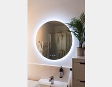Modernes Badezimmer mit einem runden, hinterleuchteten Spiegel über einem Waschbecken. Im Spiegel wird eine verglaste Duschkabine reflektiert, daneben ein weißer Heizkörper; im Vordergrund eine Kommode mit einer Zimmerpflanze.