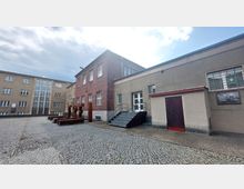 Innenhof eines historischen Gebäudekomplexes mit Backsteinfassade und verputzten Wänden. Es sind Gitterfenster, ein Zugang mit Treppe und ein Kunstwerk aus Metallbuchstaben mit dem Wort "FREI" zu sehen, der Boden ist mit Kopfsteinpflaster bedeckt.