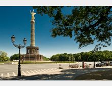 Die Siegessäule in Berlin, eine hohe Säule mit der vergoldeten Statue der Viktoria an der Spitze, steht auf einem runden Sockel mit Säulen. Im Vordergrund sind eine Straße mit Straßenlaternen und parkende Fahrräder, im Hintergrund Bäume und blauer Himmel.