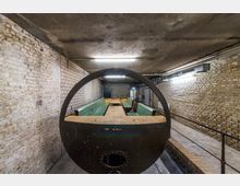 Industrieähnlicher Kellerraum mit freiliegenden Ziegelwänden und einem alten metallenem Braukessel im Vordergrund. Im Hintergrund sind grüne Holzelemente sowie eine schlichte Beleuchtung an der Betondecke sichtbar.