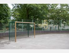 Ein kleiner Fußballplatz mit einem Metalltor, umgeben von einem hohen Zaun. Im Hintergrund sind Bäume, Wohngebäude und ein Spielplatz zu sehen.