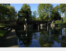 Wasserwehr in einer Flusslandschaft, umgeben von dichtem Grün und Bäumen; sichtbare Betonstruktur mit Metallgeländer, Treppen und einer kleinen Brücke. Im Vordergrund reflektiert das ruhige Wasser die umliegende Natur.
