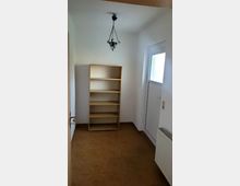 Kleiner Raum mit weißen Wänden, braunen Fliesenboden und einer hölzernen Regalwand. Im Raum befinden sich eine einfache Hängelampe an der Decke, eine Tür mit Milchglas, und ein Heizkörper an der rechten Wand.