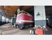 Innenansicht eines historischen Lokschuppens mit einer rot-beigen Diesellokomotive im Vordergrund, umgeben von Werkbänken und Werkzeugen. Der Raum hat eine Holzdecke, große Fenster und eine industrielle Atmosphäre.