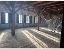 Ein rustikaler Dachboden mit Holzbalken, Holzdielenboden und mehreren großen, halbrunden Fenstern, durch die Sonnenlicht einfällt. Im Raum befindet sich eine kunstvolle Netzinstallation sowie eine angelehnte Holzleiter.