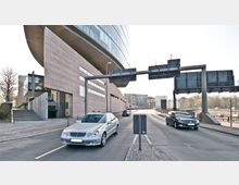 Straßenansicht mit zwei Pkw auf einer zweispurigen Fahrbahn vor einem modernen Gebäude mit geschwungener Fassade und Beton- sowie Glas-Elementen. Im Vordergrund eine große Verkehrsüberführung mit Schilderträgern, im Hintergrund Bäume und mehrstöckige Wohngebäude.