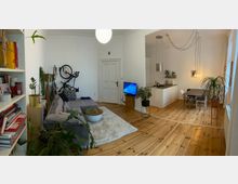 Wohnzimmer mit offener Küche in einer hellen Wohnung, ausgestattet mit Holzdielenboden und weißen Wänden. Links ein Bücherregal und eine graue Couch mit Pflanzen, rechts eine moderne Küche mit Esstisch und Hängelampen, Fahrrad an der Wand montiert.