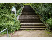 Breite Steintreppe in einer bewaldeten Umgebung, flankiert von grünen Geländern und dichtem Pflanzenbewuchs; links steht ein Schild mit Wanderhinweisen.