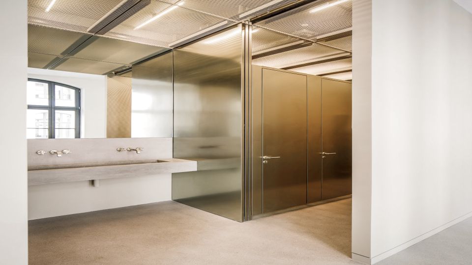 Moderner Sanitärbereich mit Betonwaschrinne und mehreren Armaturen vor einer metallverkleideten Trennwand. Rechts sind Kabinentüren in bronzefarbenen Metallpaneelen, darüber eine Rasterdecke mit linearen Leuchten und links ein Fenster.