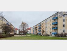 Wohnsiedlung mit mehrstöckigen Gebäuden in heller Fassade und Balkonen mit blauen Geländern. In der Mitte befindet sich eine begrünte Freifläche mit einem kleinen Spielplatz und umliegenden Bäumen.