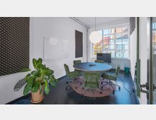 Ein moderner Besprechungsraum mit einem runden Tisch, umgeben von grünen Bürostühlen auf einem Teppich. Die weißen Wände sind mit Akustikpaneelen und einem Whiteboard versehen, und große Fenster lassen Tageslicht herein, während im Vordergrund eine grüne Zimmerpflanze steht.