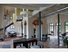 Innenraum eines modernen Cafés oder Veranstaltungsraums mit hohen Decken, weißen Wänden und freiliegenden Stahlträgern. Der Raum verfügt über große Fenster, Backsteinböden, schlichte Holzmöbel, eine Metalltreppe mit Zwischenebene und helle, zylindrische Deckenlampen.